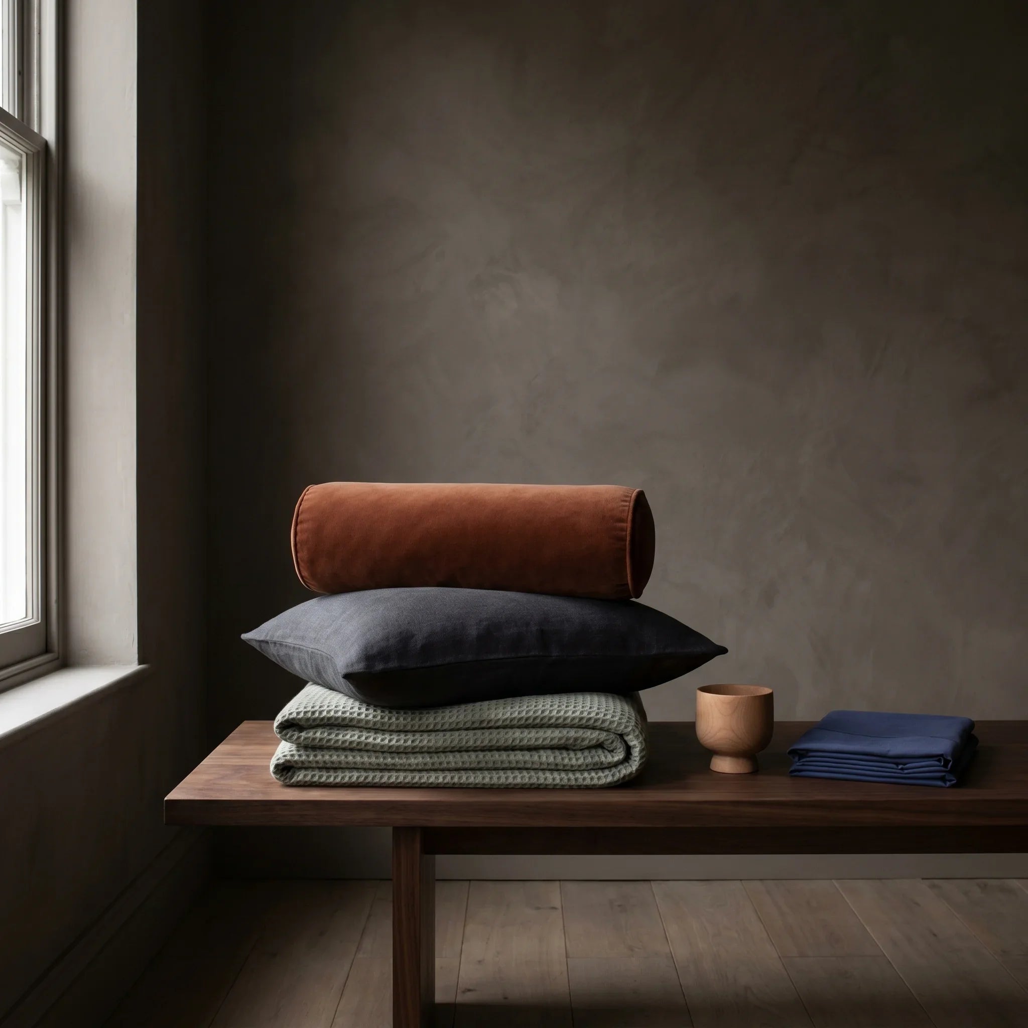 All Wellnatura products: a sage waffle throw, charcoal linen cushion, rust velvet bolster, beech wood vessel and indigo bamboo pillowcase arranged on a walnut table